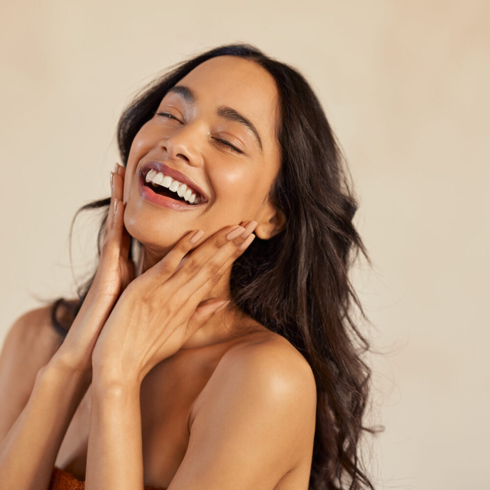 Multiethnic young woman touching her clean and healthy face against background with closed eyes. Latin hispanic woman feeling good with perfet skincare routine. Natural beauty portrait of mexican girl with black long hair.